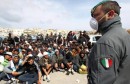 epa02659783 A masked police officer  stands over a large group of migrants who are gathered together for identification ourposes in the port area of  Lampedusa,  March 29 2011. The Sicily Regional Government said there are 6,200 migrants on the island. Six ships with a total capacity of 10,000 berths are to take migrants from the island tomorrow,  according to the extraordinary commissioner for the humanitarian emergency  EPA/VENEZIA FILIPPO