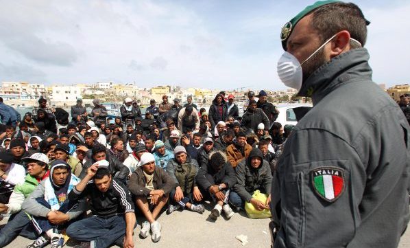 epa02659783 A masked police officer  stands over a large group of migrants who are gathered together for identification ourposes in the port area of  Lampedusa,  March 29 2011. The Sicily Regional Government said there are 6,200 migrants on the island. Six ships with a total capacity of 10,000 berths are to take migrants from the island tomorrow,  according to the extraordinary commissioner for the humanitarian emergency  EPA/VENEZIA FILIPPO