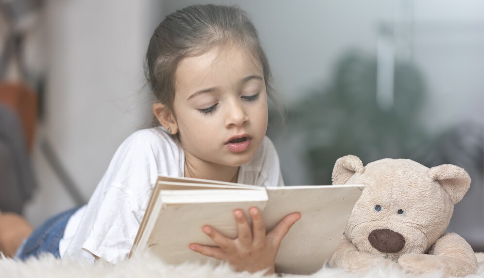 portrait-cute-little-girl-reading-book-home-lying-floor-with-her-favorite-toy_169016-13795